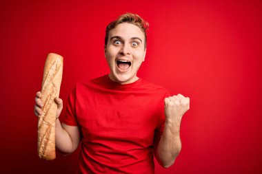 Genç ve yakışıklı kızıl saçlı adam izole edilmiş kırmızı arka planda taze ev yapımı ekmek tutuyor gururla bağırıyor, zaferi ve başarıyı kutluyor kollarını kaldırarak heyecanla bekliyor.