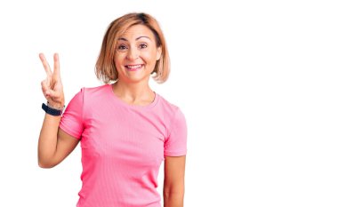 Spor kıyafetleri giyen genç sarışın kadın kendini beğenmiş ve mutlu bir şekilde gülümserken iki numarayı gösterip işaret ediyor.. 