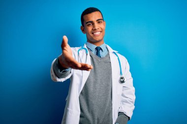 Yakışıklı Afro-Amerikan doktor ceketli ve steteskop takmış mavi arka planda gülümseyen, yardım eli uzatan ve kabul gören..