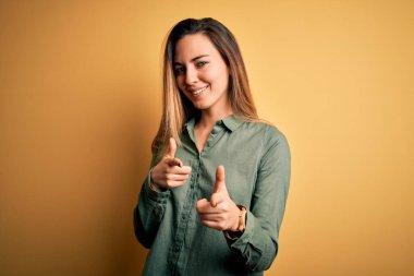 Sarı arka planda yeşil tişörtlü, mavi gözlü güzel sarışın kadın mutlu ve komik yüzlü parmaklarını kameraya doğrultuyor. İyi enerji ve titreşimler.