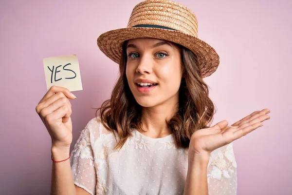 Young blonde girl wearing a hat holding paper note with yes word as positive message very happy and excited, winner expression celebrating victory screaming with big smile and raised hands