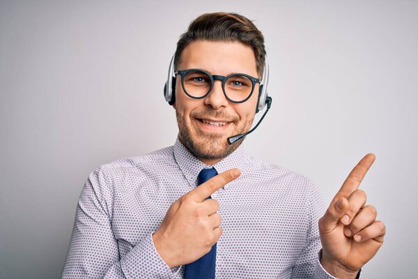 Young call center operator business man with blue eyes wearing glasses and headset smiling and looking at the camera pointing with two hands and fingers to the side.