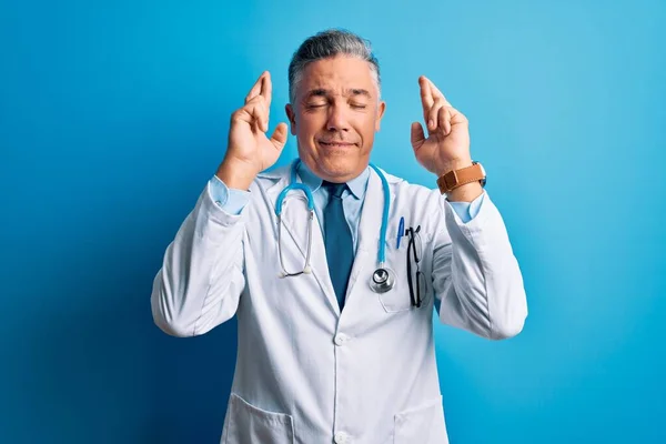 中年英俊的白发苍苍的医生 身穿外套 戴着蓝色听诊器 摆弄着手指 带着希望微笑 闭上了眼睛 运气和迷信观念 — 图库照片