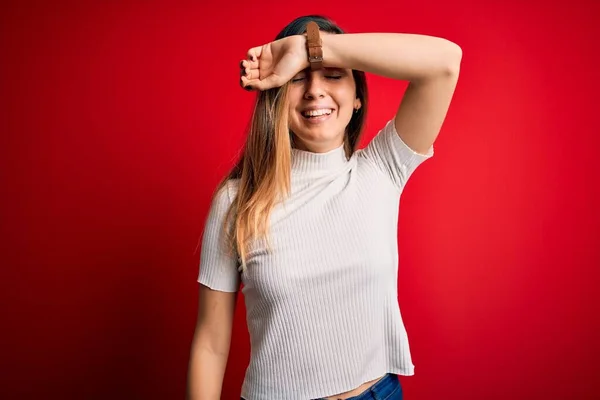 Beautiful blonde woman with blue eyes wearing casual white t-shirt over red background covering eyes with arm smiling cheerful and funny. Blind concept.