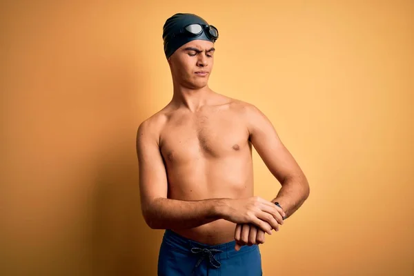 年轻英俊的男子穿着泳衣 戴着泳帽 披着孤立的黄色背景 用腕表查看时间 放松而自信 — 图库照片