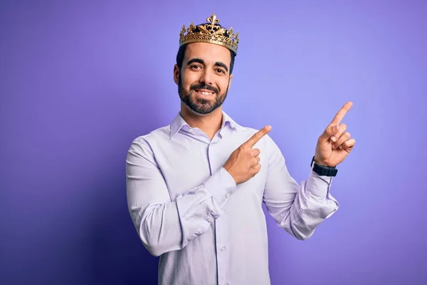 年轻英俊的男人留着胡子 头戴金色的王冠 背景是紫色的 他笑着看着摄像机 用双手和手指指向旁边 — 图库照片