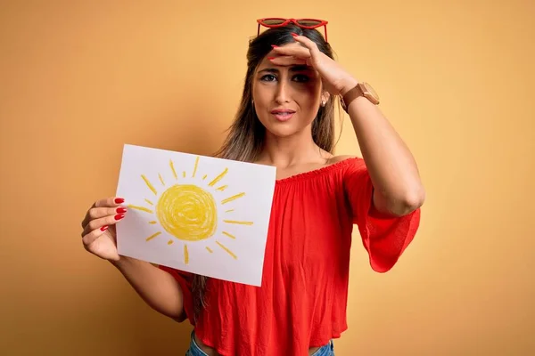 Young beautiful brunette woman holding paper with sun draw over isolated yellow background stressed with hand on head, shocked with shame and surprise face, angry and frustrated. Fear and upset for mistake.