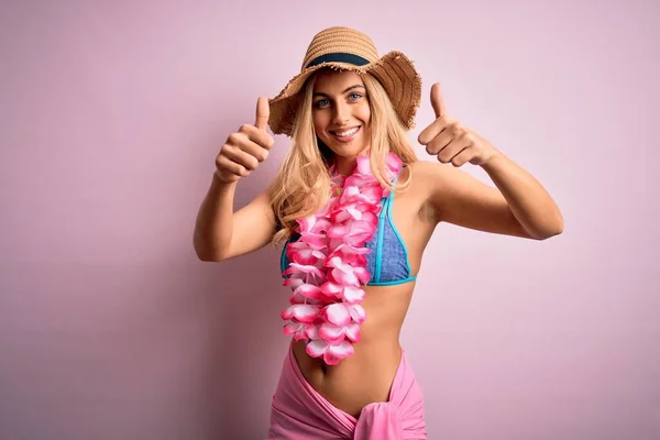 Young beautiful blonde woman on vacation wearing bikini and hat with hawaiian lei flowers approving doing positive gesture with hand, thumbs up smiling and happy for success. Winner gesture.