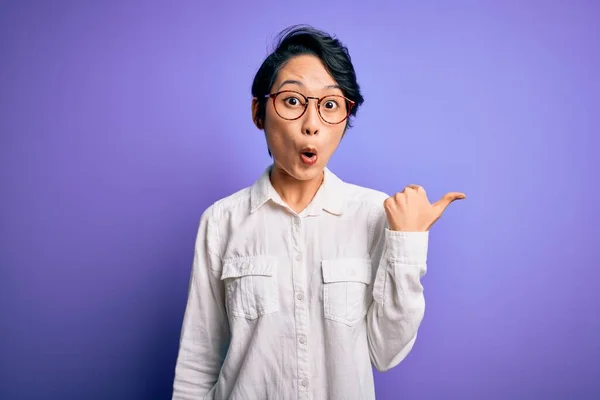 Young beautiful asian girl wearing casual shirt and glasses standing over purple background Surprised pointing with hand finger to the side, open mouth amazed expression.