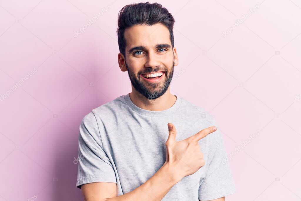 Young handsome man with beard wearing casual t-shirt smiling cheerful pointing with hand and finger up to the side 