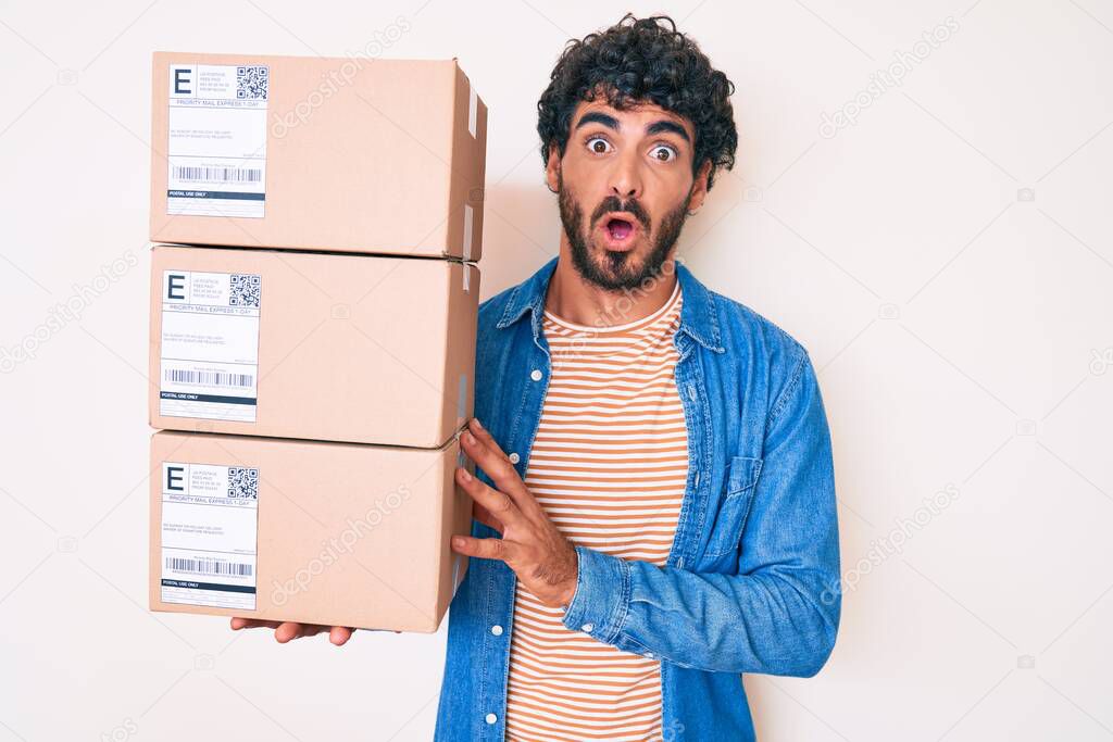 Handsome young man with curly hair and bear holding delivery package scared and amazed with open mouth for surprise, disbelief face 