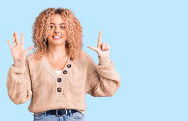 Kış kazağı giyen, kıvırcık saçlı genç sarışın kadın yedi numaralı parmaklarıyla kendini beğenmiş ve mutlu gülümserken kendini gösteriyor.. 