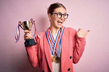 Genç, güzel, kızıl saçlı, elinde madalyalarla pembe arka planı işaret eden ve gülümseyen bir yüzü olan bir kadın.