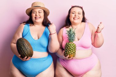 Genç ve büyük beden ikizler ellerinde kavun ve ananas tutuyorlar gururla bağırıyorlar zaferi kutluyorlar ve başarıyı kollarını kaldırarak kutluyorlar. 
