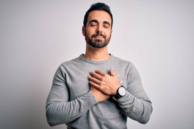 Sakallı, sıradan bir süveter giyen, beyaz arka plan üzerinde duran, elleri göğsünde, gözleri kapalı ve yüzünde minnet ifadesi olan genç yakışıklı bir adam. Sağlık kavramı.