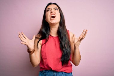 Genç esmer kadın pembe izole edilmiş arka plan üzerine gündelik bir tişört giyiyor deli gibi bağırıyor ve bağırıyor agresif bir ifade ve kollar havada. Hayal kırıklığı konsepti.