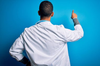 Yakışıklı Afrikalı Amerikalı doktor ceketli ve steteskop takmış mavi arka planda geriye doğru poz veriyor.