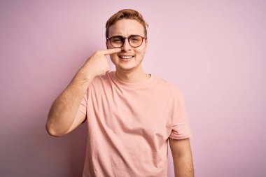 Genç, kızıl saçlı, günlük tişört giyen, pembe arka plan üzerinde duran, el parmağı ile yüzünü ve burnunu gösteren, neşeli gülümseyen bir adam. Güzellik konsepti