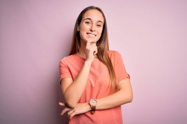 Izole pembe arka plan üzerinde duran rahat t-shirt giyen genç güzel kadın çapraz kolları ve çene üzerinde kaldırdı el ile gülümseme ile kameraya emin bakıyor. Olumlu düşünme.