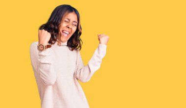 Genç, güzel, melez, balıkçı yaka kazak giyen, mutlu ve heyecanlı genç bir kadın kolları havada, gülümseyen ve başarı için çığlık atan, kazanan bir jest yapıyor. kutlama konsepti. 