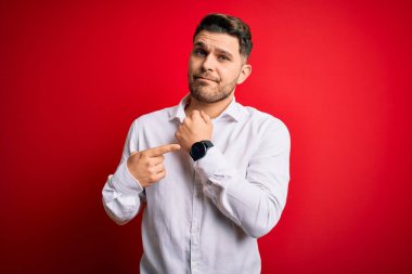 Mavi gözlü, zarif gömlekli genç bir iş adamı kırmızı izole arka planda dikiliyor. Aceleyle saati, sabırsızlığı ve rahat bir ifadeyle kameraya bakıyor.