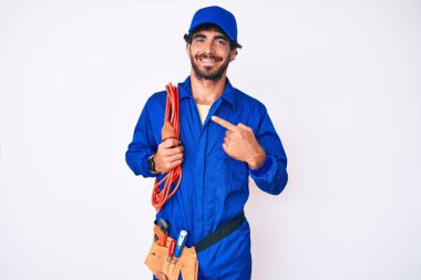 Kıvırcık saçlı, ayı kılıklı, işçi tulumu giyen ve el ve parmaklarıyla mutlu bir şekilde gülümseyen yakışıklı bir genç. 