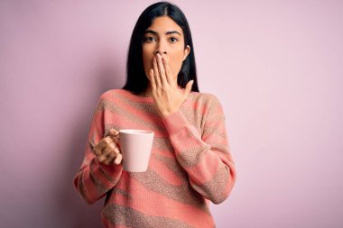 Genç, güzel İspanyol bir kadın pembe izole edilmiş arka planda bir fincan sıcak kahve içiyor. Ağzı kapalı, utancından ağzı açık, korkunun ifadesi, sessizlikten korkmak, gizli bir konsept.
