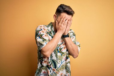 Tatilde mavi gözlü genç adam sarı arka planda çiçekli bir tişört giyiyor ve ağlarken suratını ellerle kapatıyor. Depresyon kavramı.