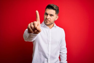 Mavi gözlü, zarif gömlekli genç bir iş adamı. Kırmızı izole edilmiş arka planda duruyor. Parmağını kaldırmış ve kızgın bir ifadeyle işaret ediyor.