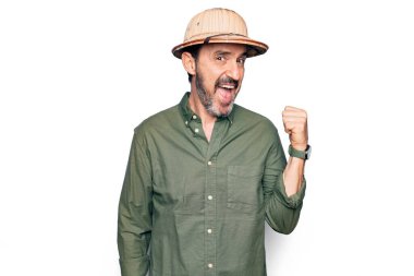 Tatilde olan orta yaşlı bir turist izole edilmiş beyaz arka plan üzerinde kaşif şapkası takıyor başparmağını yana uzatıyor ve ağzı açık gülümsüyor.