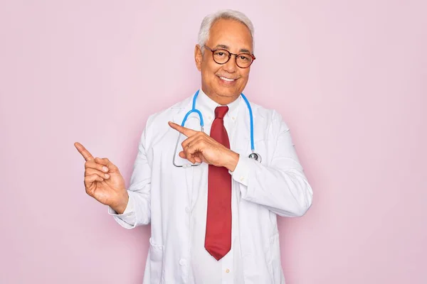 中年シニア白髪の医師男を身に着けています聴診器とプロの医療コート笑顔とカメラを見てポインティングで二本の手と指で側面 — ストック写真