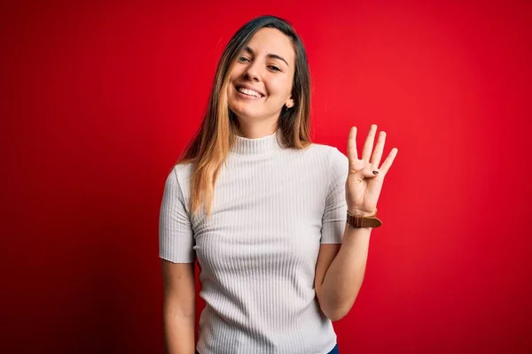 Beautiful blonde woman with blue eyes wearing casual white t-shirt over red background showing and pointing up with fingers number four while smiling confident and happy.