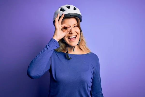 Young beautiful blonde cyclist woman wearing bike security helmet over purple background doing ok gesture with hand smiling, eye looking through fingers with happy face.