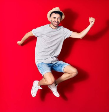 Genç, yakışıklı, günlük kıyafetler giyen ve şapkalı mutlu bir İspanyol. Yüzünde tebessümle zıplıyor, kırmızı arka planda yumruklar havada kutluyor..