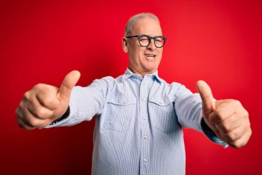 Orta yaşlı, beyaz, sıradan çizgili gömlek giyen ve kırmızı arka planda gözlük takan yakışıklı bir adam elinle olumlu jest yapmayı onaylıyor, gülümsüyor ve başarı için seviniyor. Kazanan jest.