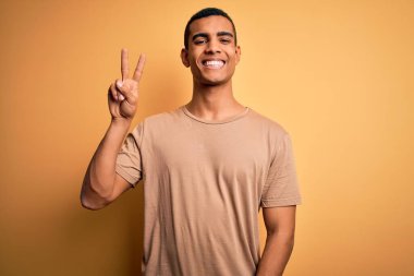 Genç, yakışıklı, Afro-Amerikalı bir adam sarı arka planda dikilen günlük tişörtler giyer ve iki numarayı gösterirken kendinden emin ve mutlu bir şekilde gülümserdi..