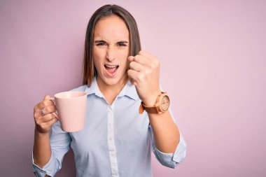 Genç, güzel bir kadın, izole edilmiş pembe arka planda dikilmiş kahve içerken sinirlenmiş ve sinirlenmiş bir şekilde bağırıyor, çıldırmış ve el kaldırarak bağırıyor. Öfke kavramı.