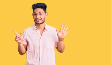 Yakışıklı Latin Amerikalı rahat bir yaz tişörtü giyen genç adam yedinci parmaklarını gösterip gösterirken kendinden emin ve mutlu gülümsüyor.. 