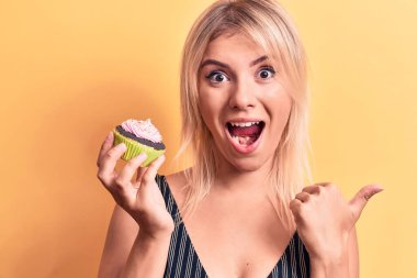 Sarışın, güzel bir kadın, izole edilmiş sarı arka planda tatlı çikolatalı kek yiyor. Başparmağıyla yan tarafa bakıyor, mutlu bir şekilde gülümsüyor.