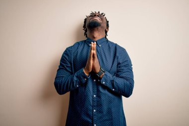 Genç, yakışıklı, Afro-Amerikan bir adam, günlük gömleğini giymiş beyaz arka plan üzerinde duruyor, yalvarıyor ve dua ediyor. Yüzünde umut ifadesi olan çok duygusal ve endişeli bir ifade var. Yalvarıyorum..