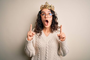 Kıvırcık saçlı, beyaz arka planda altın kraliçenin tacını takan genç ve güzel bir kadın şaşırmış ve şaşırmış bir şekilde parmaklarıyla işaret ediyor ve kollarını kaldırıyordu..