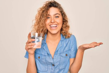 Mavi gözlü genç sarışın kadın beyaz arka planda bir bardak su içiyor çok mutlu ve heyecanlı, kazanan yüz ifadesi zaferi büyük bir gülümsemeyle kutluyor ve ellerini kaldırıyor.