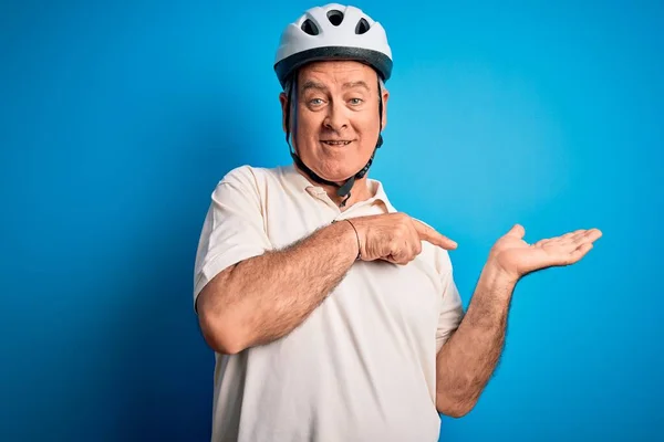 Middle age hoary cyclist man wearing bike security helmet over isolated blue background amazed and smiling to the camera while presenting with hand and pointing with finger.