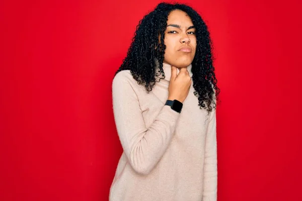 Young african american curly woman wearing casual turtleneck sweater over red background Touching painful neck, sore throat for flu, clod and infection