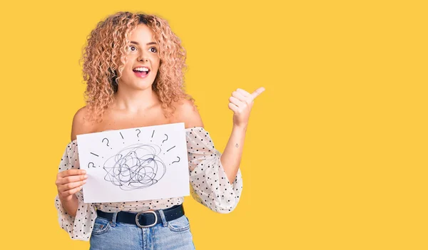 Young blonde woman with curly hair holding scribble draw pointing thumb up to the side smiling happy with open mouth