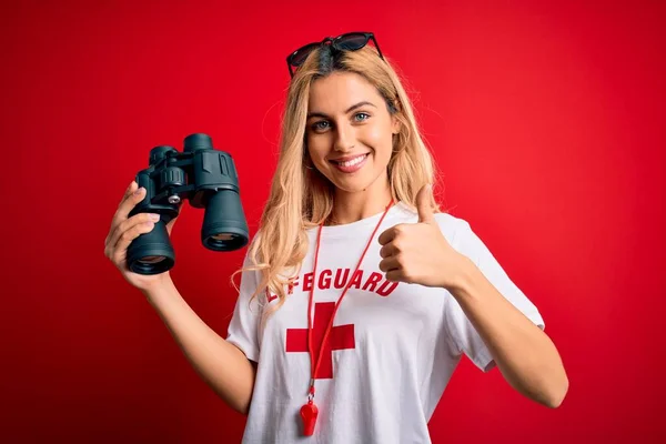 Young beautiful blonde lifeguard woman wearing t-shirt with red cross using binoculars happy with big smile doing ok sign, thumb up with fingers, excellent sign