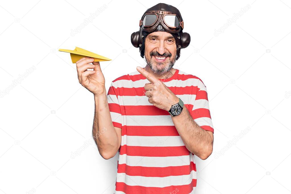 Middle age aviator man wearing vintage helmet and glasses holding paper airplane smiling happy pointing with hand and finger