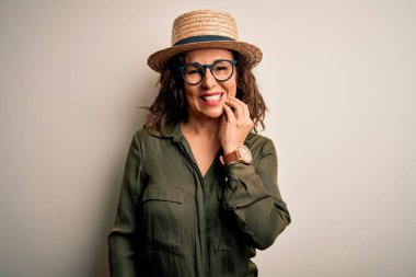 Orta yaşlı esmer kadın, gözlüklü ve şapkalı, izole edilmiş beyaz arka planda duruyor. Diş ağrısı ya da diş hastalığından dolayı acı dolu bir ifadeyle ağıza dokunuyor. Dişçi