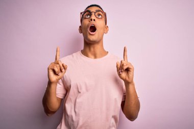 Yakışıklı Afro-Amerikan erkeği pembe arka planda gündelik tişört ve gözlük takıp şaşkın şaşkın yukarı bakıyor, parmaklarıyla işaret ediyor ve kollarını kaldırıyordu..