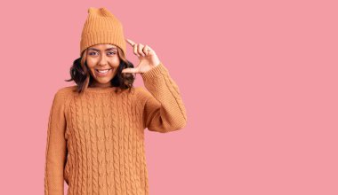 Genç, güzel, melez, yün kazak giyen, şapkalı, gülümseyen, kendine güvenen, el hareketi yapan, parmaklarıyla işaret yapan ve kameraya bakan bir kadın. ölçüm kavramı. 